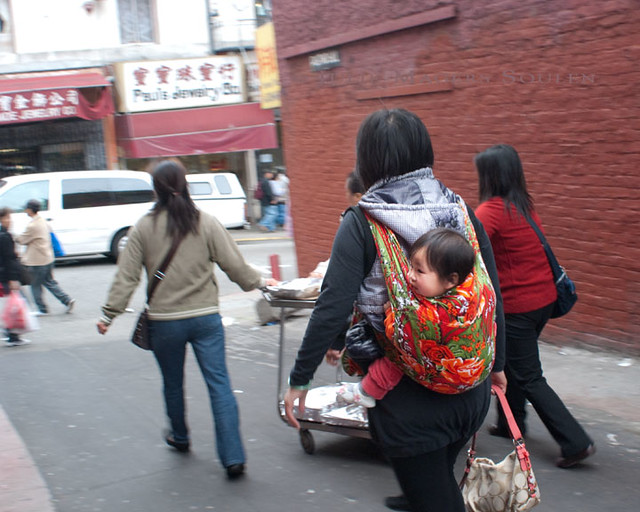 A group of women push a cart of food down an alley in Chinatown, one with a baby strapped to her back.