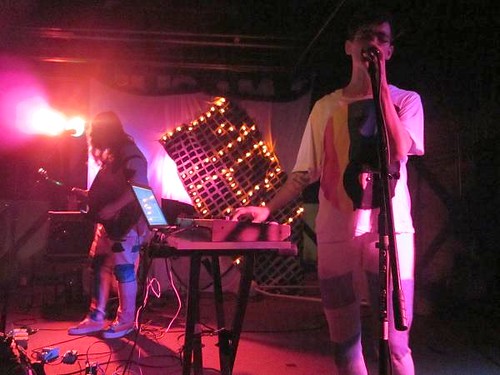 JD Samson is on stage singing into a mic and wearing a t-shirt with a large rainbow yin-yang on it. Bassist Tammy Hart can be seen in the background