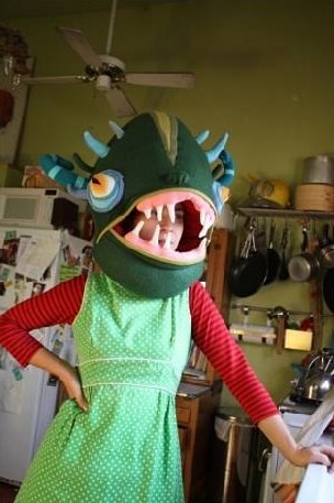 Jennifer Strunge, a white woman in a long-sleeved red shirt under a sleeveless green dress, stands with her right hand on her hip. She has one of her monster creatures over her entire head, and it looks like the head of a vicious dark green fish. The creature has four little light blue horns sticking from its top, two on each side, above blue-rimmed eyes with yellow irises. Jennifer's head is visible through its mouth, which is an opening, between two rows of jagged, sparse white teeth.