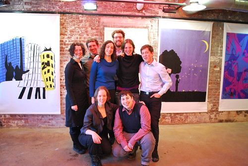 group shot of 8 alumni, posing for the camera, with a brick wall with some large paintings in the background