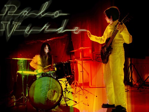 The two women of Palo Verde wear yellow suits. One plays a guitar and one is behind a drum kit. 