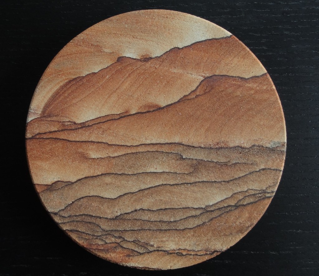 Image shows a round coaster on a black wood background. It's variations of brown, russet, and tan. It looks like someone's painted a mountainous desert landscape complete with foothills on it, but it's all natural.