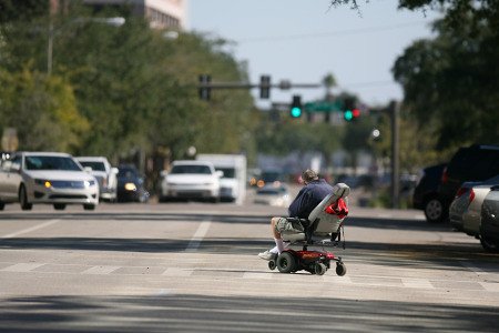 a person in a power chair crosses a pedestrian crossing on a tree lined street. our of focus cars are visible in the background, at a traffic light.