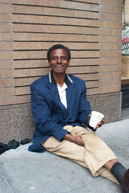 A homeless man in San Francisco smiles big for the camera as he sits on the sidewalk begging.