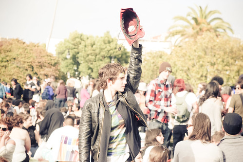 Androgynous pale person wearing a leather jacket and a t-shirt, standing in a crowd and holding up a hand with a baseball glove on it.