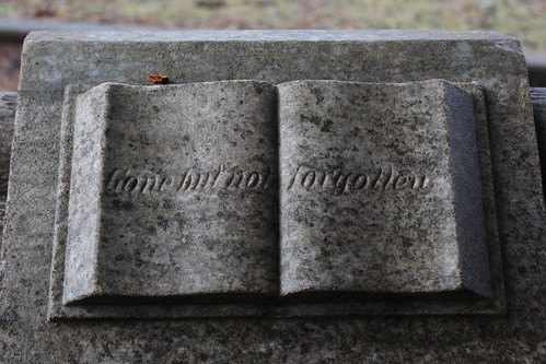 A detail of the top of a headstone. An open book has been carved as an ornament, and text across the pages reads 'gone but not forgotten'