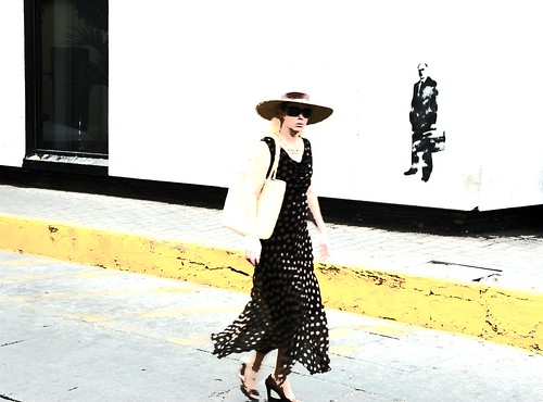 Simple: blonde Spanish woman tourist in a large straw hat, wearing a heavy gold link chain, silk dress, straw bag, guerrilla wall art of a business man with a brief case, Historical district, Mazatlan, Mexico by Wonderlane