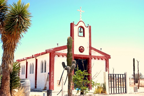 Rural white and red painted Christian church, with bell, cross of light bulbs, hub cap ventilation, fence, cactus with hose, broom, mop, water barrel, blue sky, Mexico by Wonderlane
