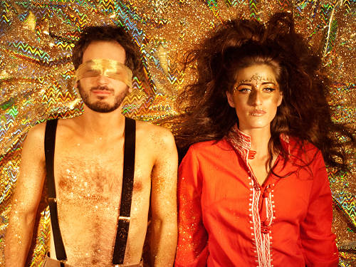 a man and a woman lie next to each other on a gold, sparkly background. the man has his eyes blindfolded with a gold piece of the cloth. the woman has her eyes open. she has a golden bronze shimmer on her face.