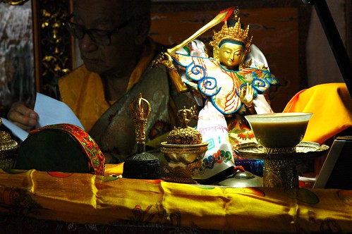 A statue of Manjushri wearing statue clothing, His Holiness Dagchen Sakya on the throne reading a text, religious items, including tea, drum, bell, skullcup, yellow ritual scarf, Tharlam Monastery of Tibetan Buddhism, Boudha, Kathmandu, Nepal by Wonderlane