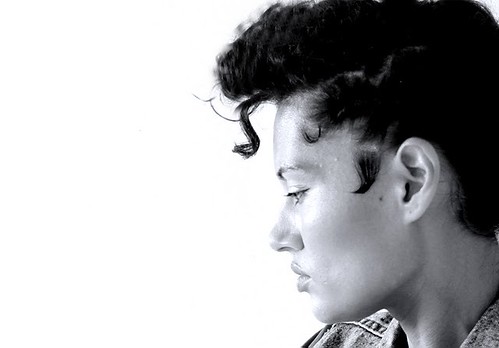 Profile of a medium-skinned person with dark, curly hair styled in an Elvis-style pompadour. They appear to be wearing a jean jacket. The photo is in black-and-white.