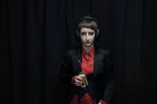 Pale-skinned androgynous person with short brown hair wearing red lipstick, a black blazer over a red shirt and black vest, and headphones. They have a half-full beer bottle in one hand.