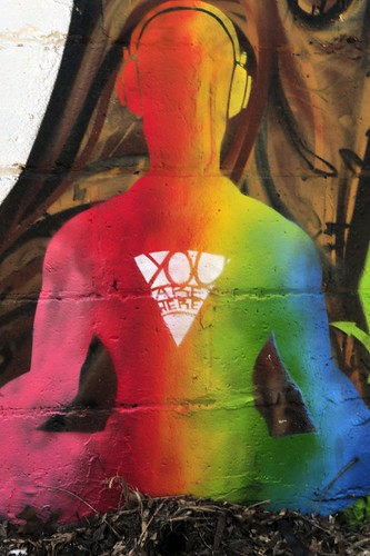 a body sitting crosslegged, wearing headphones, presented in rainbow silhouette -- over the heart is a triangle, pointed down, containing the words, 'You are here'