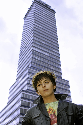 Person in leather jacket standing in front of tall building wearing a gold crown made of wire-like material.
