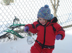 Speck in red snowsuit with play shovel in hand and grin