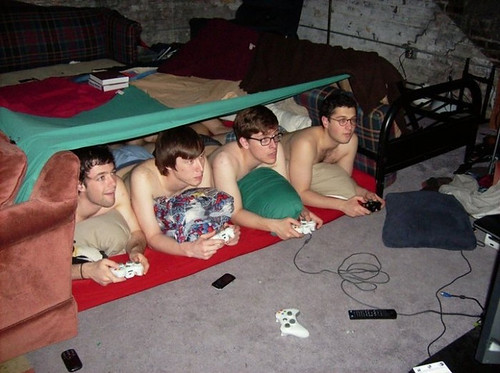 Front view of 4 people laying under a sheet tent with no shirts, playing video games in a basement