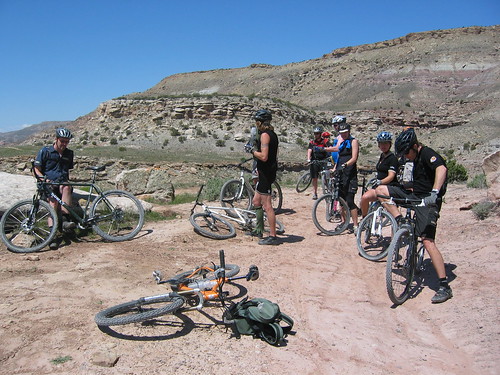 A group of cyclists and their bikes, standing on a dirt clear in the desert, with hills behind them
