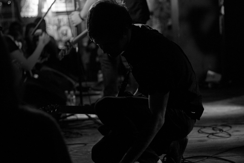Black and white concert photograph of a guitarist crouched low on stage in dramatic rim lighting during a performance.