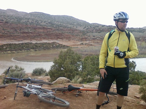 Front view of a person standing on a rock ledge with 3 bike laying near, with a river and desert hills in the background